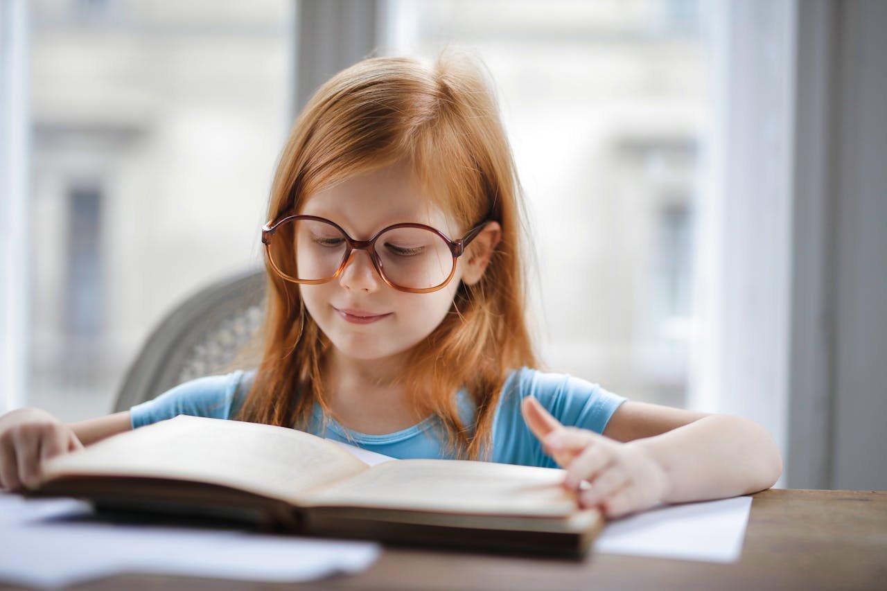 Charming young girl wearing oversized glasses reading a book at home.